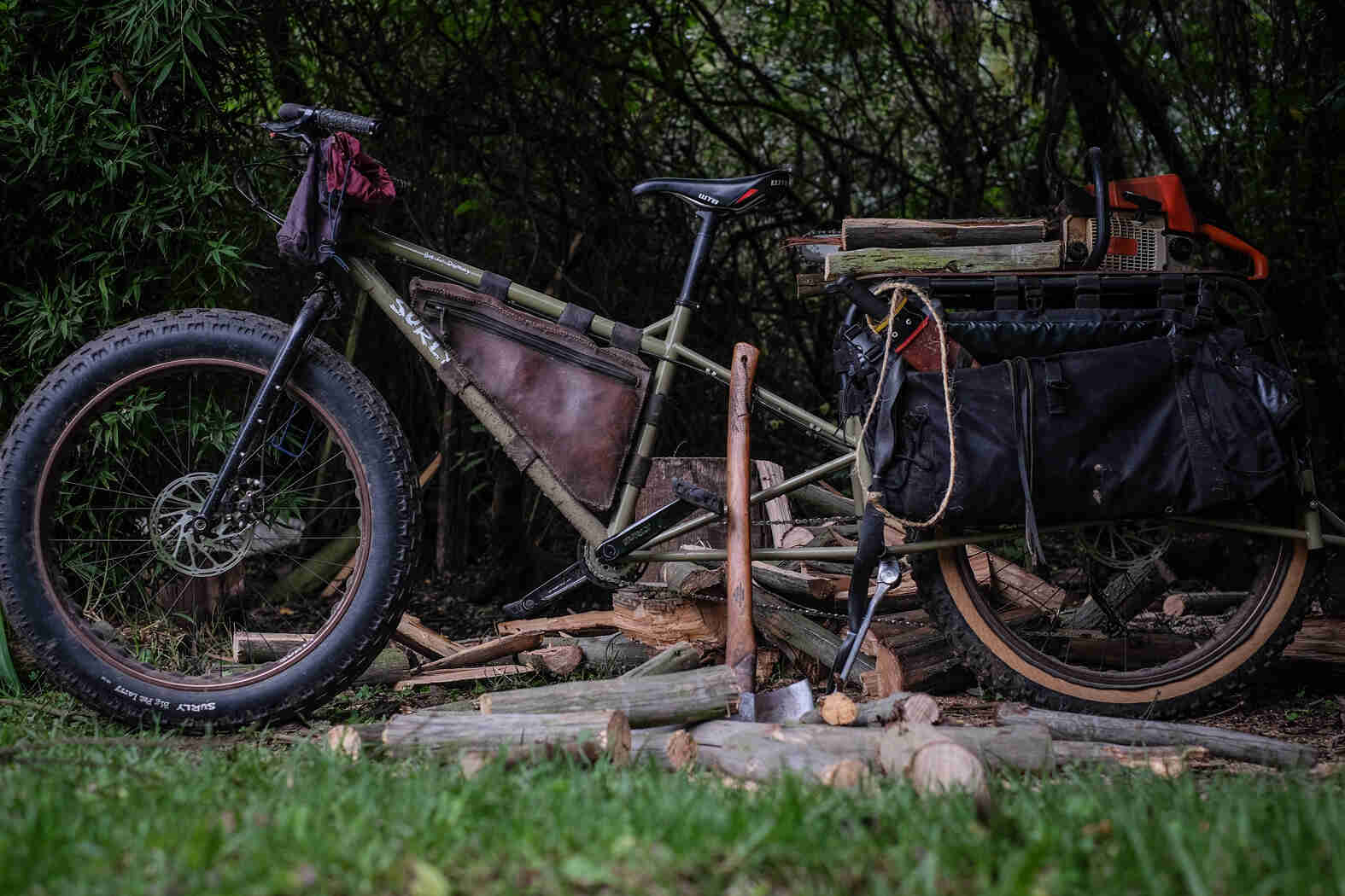 Left view of a Surly Big Fat Dummy bike, on top of axe chopped logs, with thick woods in the background