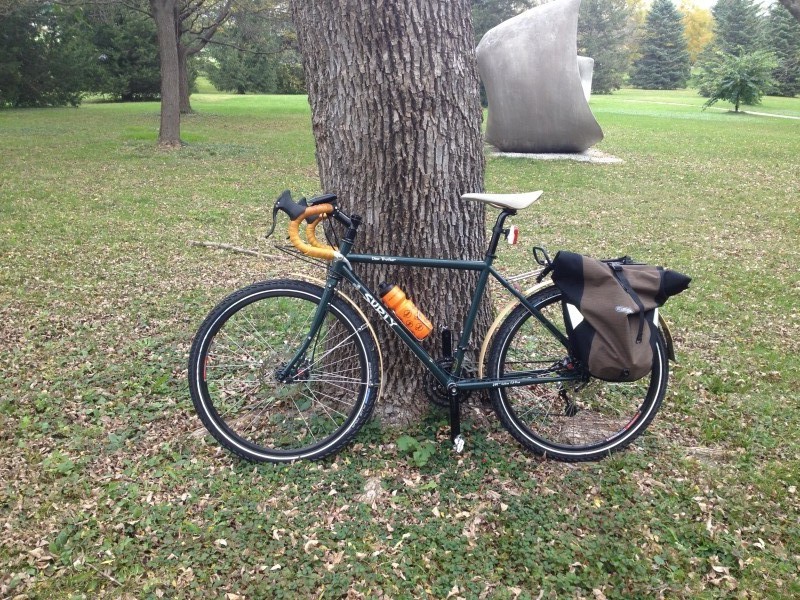 Left profile view of a Surly Disc Trucker bike, leaning on a tree in a field, with a sculpture in the background