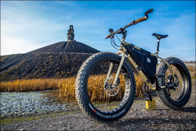 Front left angle view of a Surly fat bike on a gravel road, with a bean field and a large mount in the background