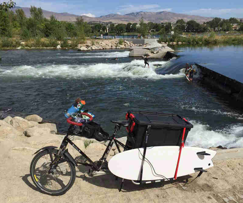 Left side view of a black Surly Big Dummy bike with a surfboard on back, on a river bank, with a surfer in the river
