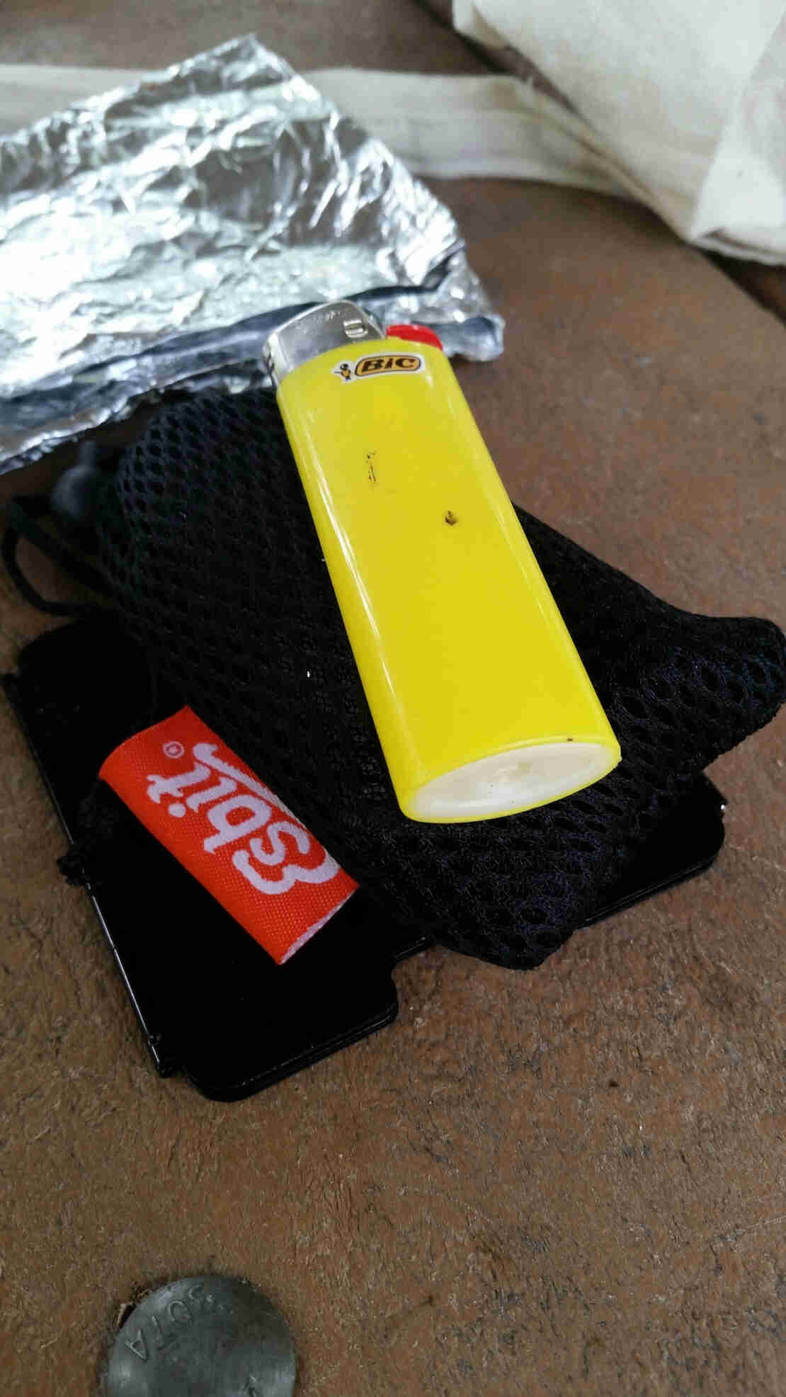 Close up view of a yellow Bic lighter on top of a black mesh sack, with foil in the background
