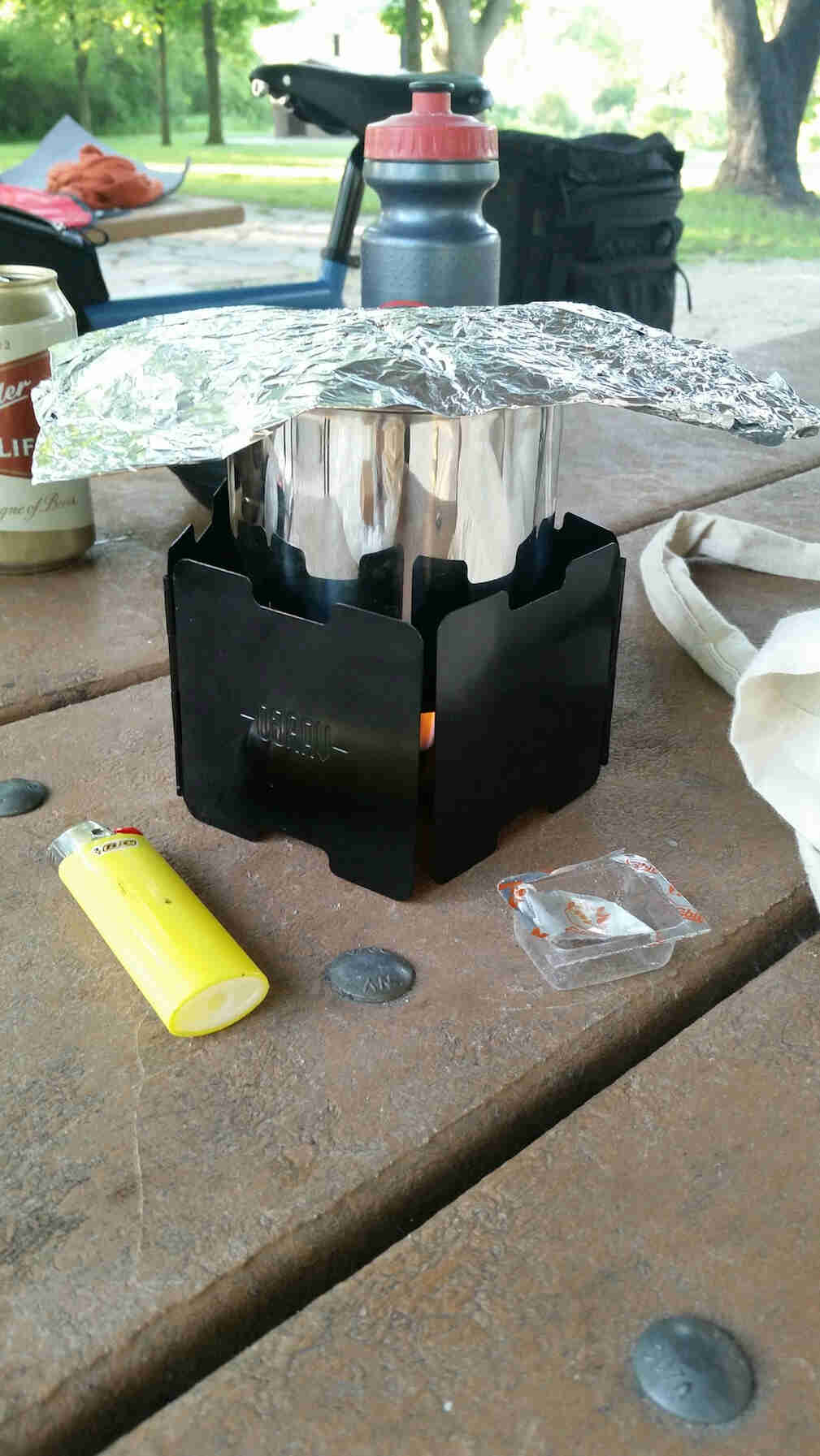 An Esbit table stove with next to a yellow Bic lighter, on top of a picnic table with a bike in the background