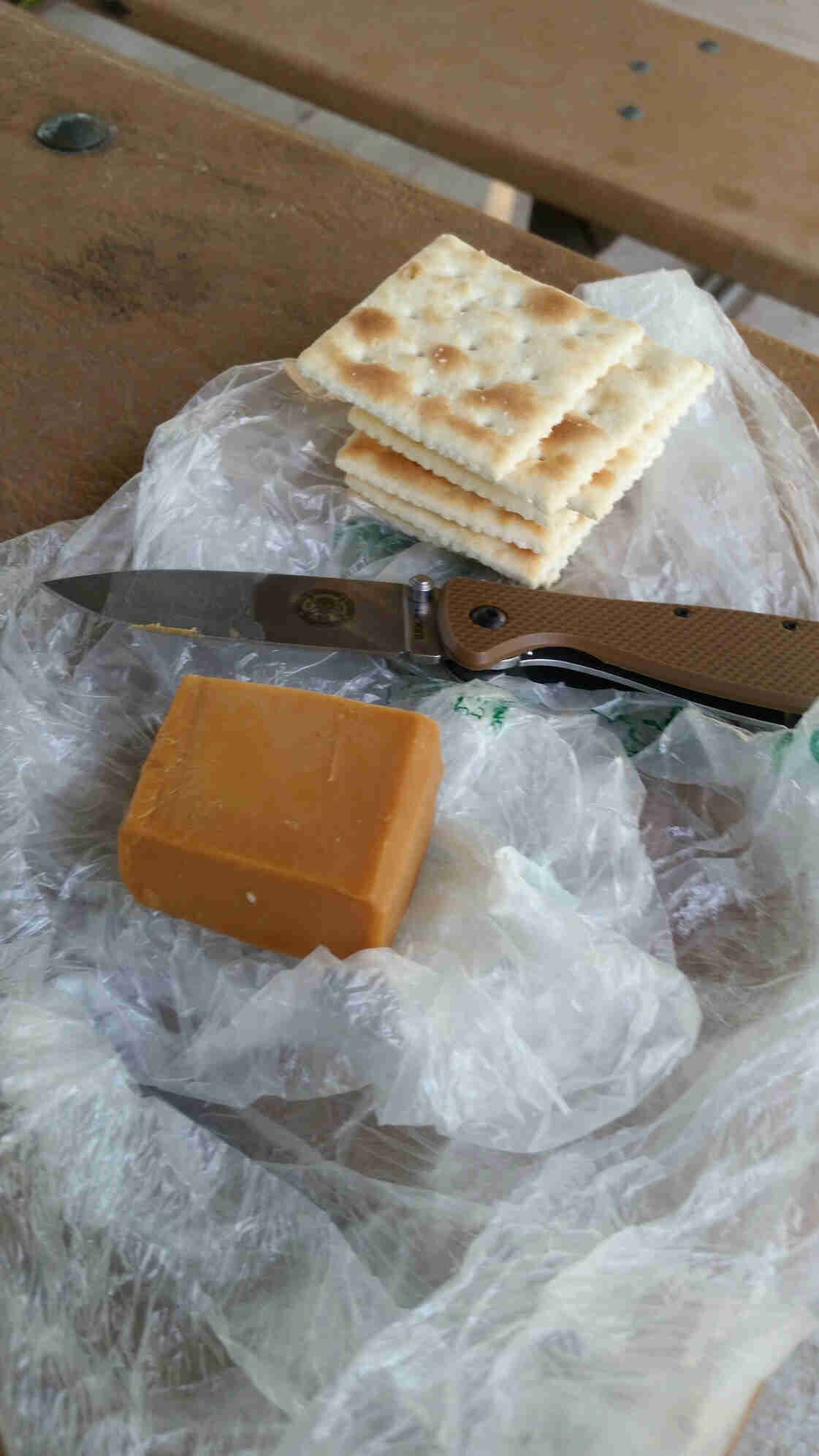 Downward view of crackers, a knife and block of cheese, on top of a clear plastic bag