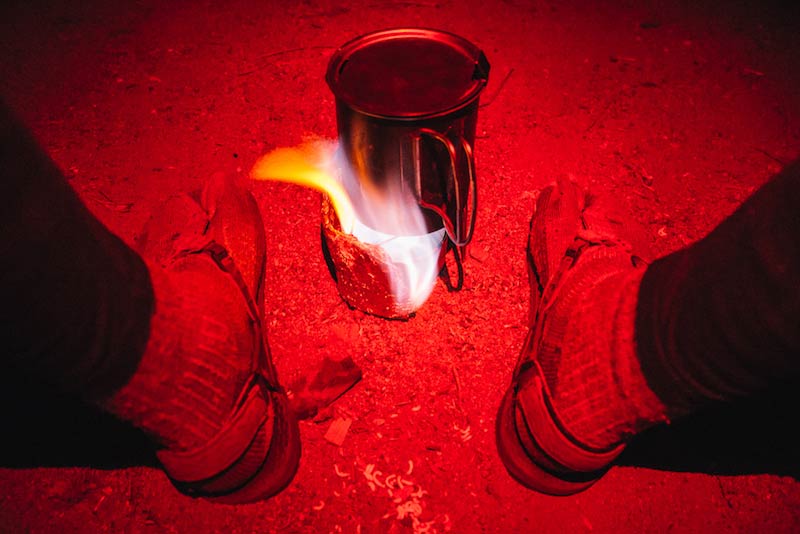 Downward view of  a steel cup with a flame underneath, between 2 sandal clad feet, and red flashing beam shining over