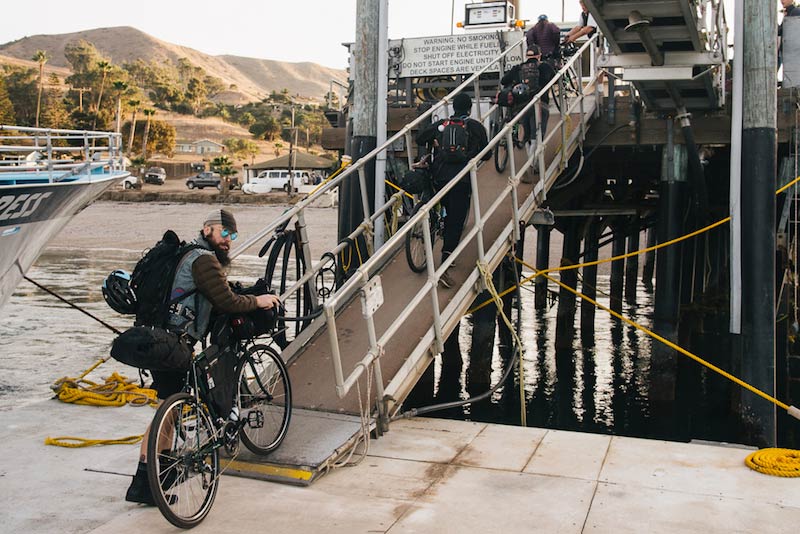 Rear view of 4 cyclists walking single file with their bikes, up a ramp at a dock, leading to a ship