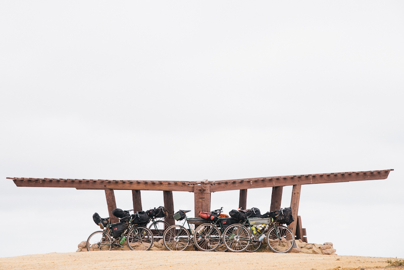 Side profiles of gear loaded bikes lined up in front of a wooden pergola on a sandy plot 