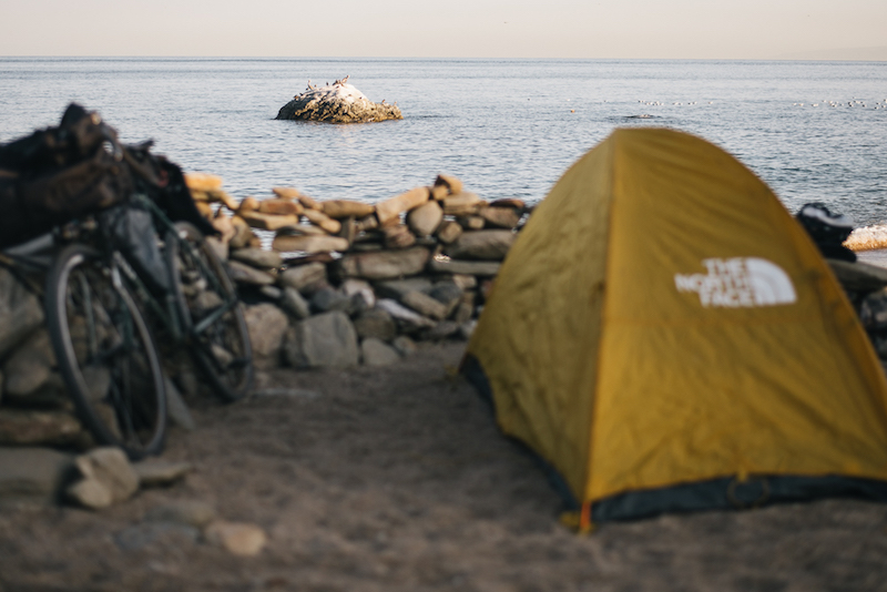 Rear view of a gear loaded bike and a yellow tent facing a rock wall with the ocean in the background