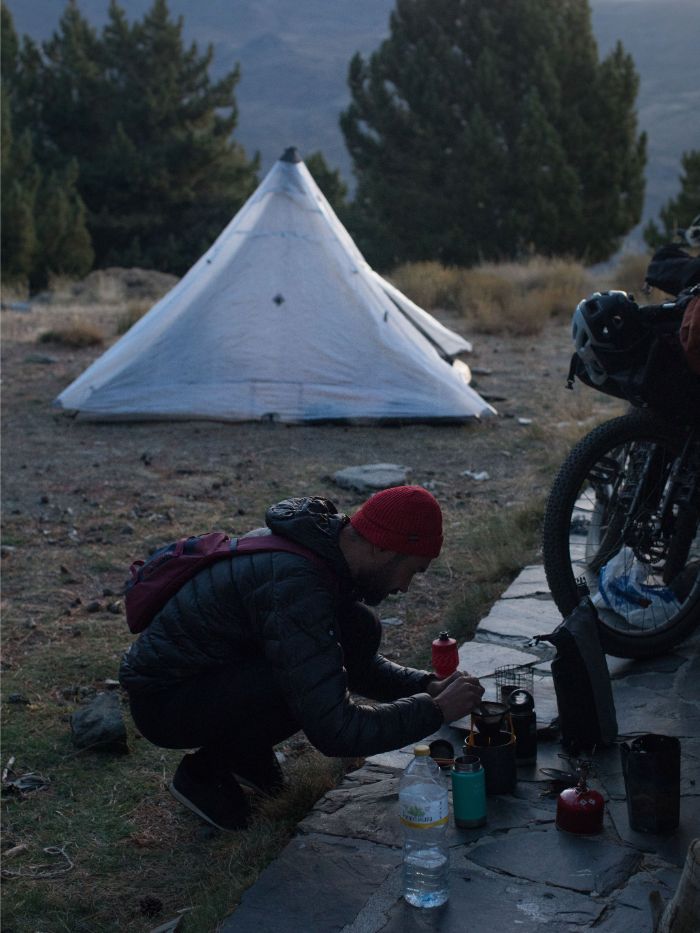 Person squatting at a campsite with a bike loaded with gear and tent with pine trees behind it in the background