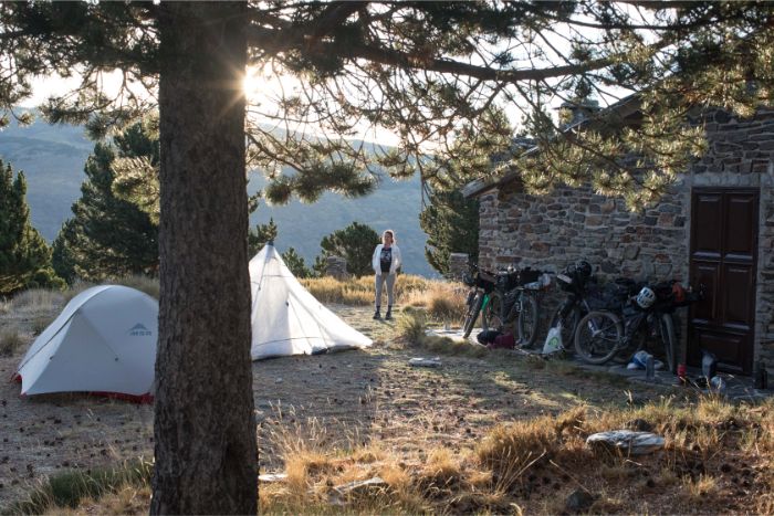 Person stands between 2 tents and a stone building with fully geared bikes leaning against it at mountain campsite