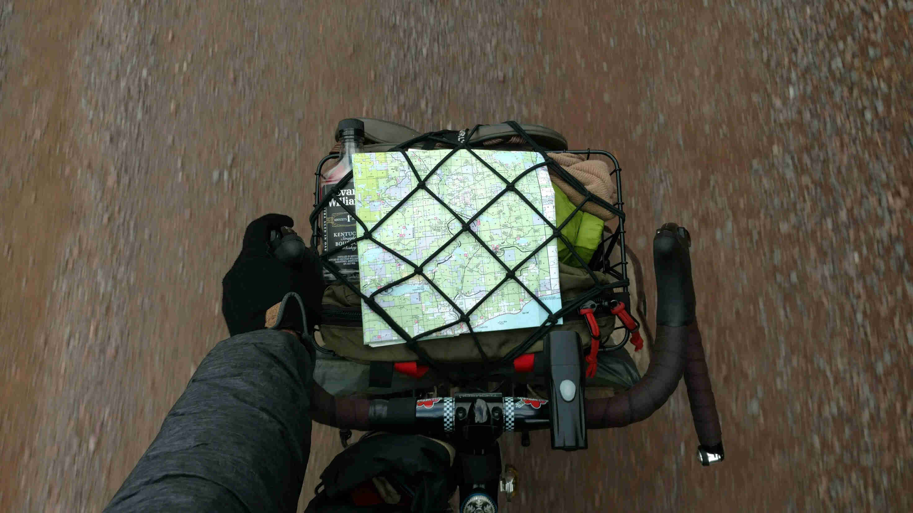 Downward view of bike handle bars and pack with a map strapped to the top, on a bike traveling down a gravel road