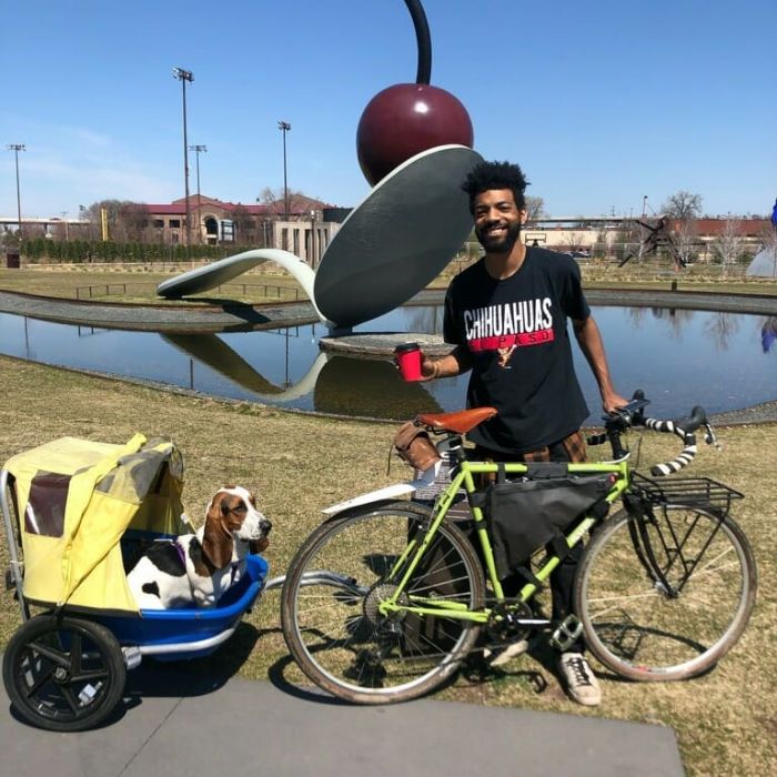 Cyclist on right side of a green bike with dog behind in a bike trailer in front of spoon and cherry sculpture
