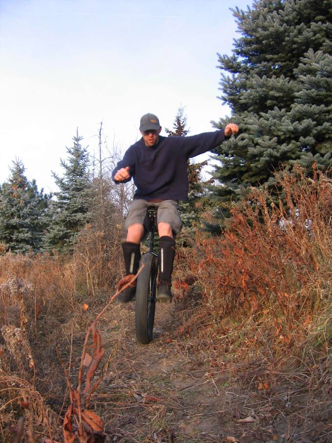 Front view of a person, riding a fat wheeled unicycle, on a dirt trail with weeds and trees on the sides