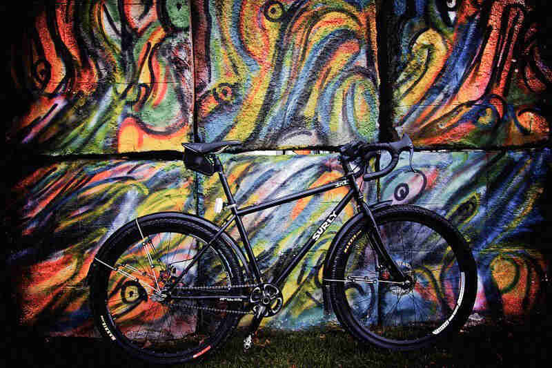 Right profile of a black Surly bike, standing in grass, against cement block wall with a colorful mural painted on it
