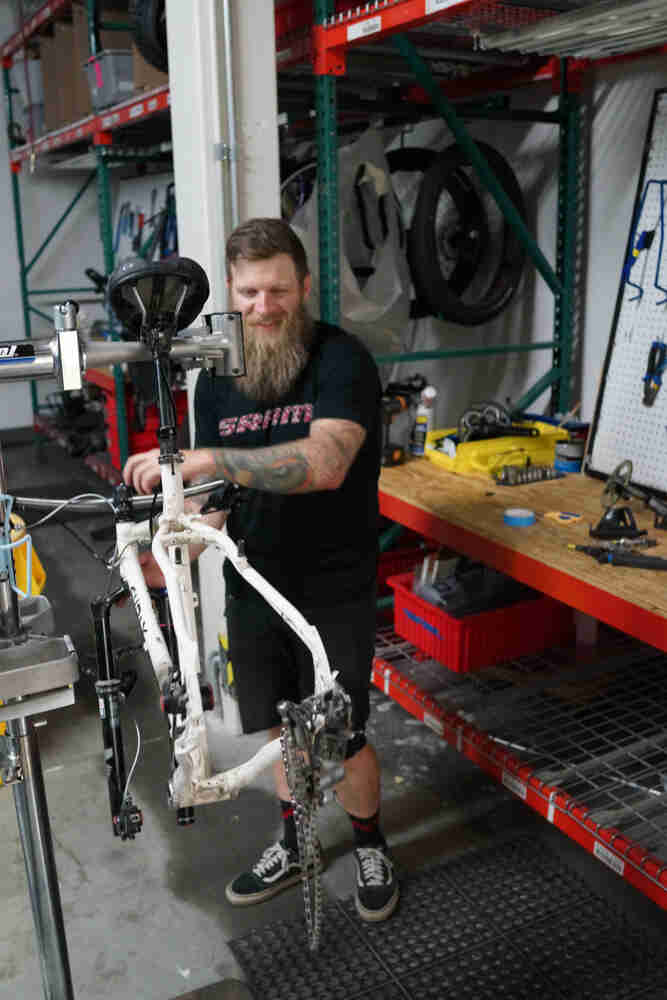 A person carrying a bike with no wheels, in a warehouse, next to a workbench