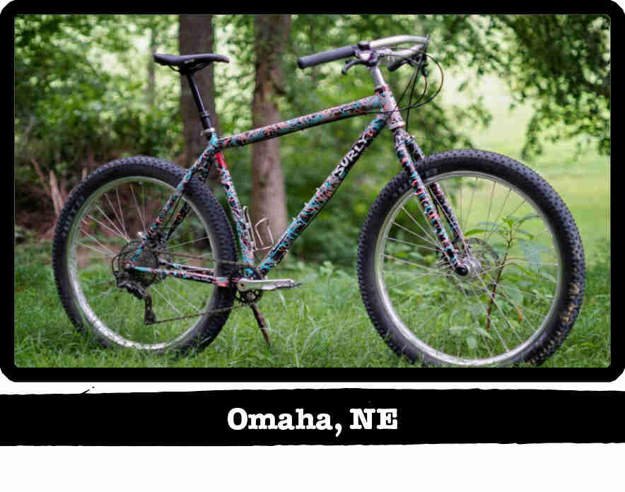 Right side view of a Surly Karate Monkey bike, multi colored, on grass with woods behind - Omaha, NE tag below image