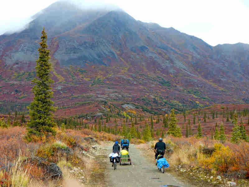 Rear view of 3 cyclists riding their bikes down a hill, on a road, with a colorful mountain ahead of them