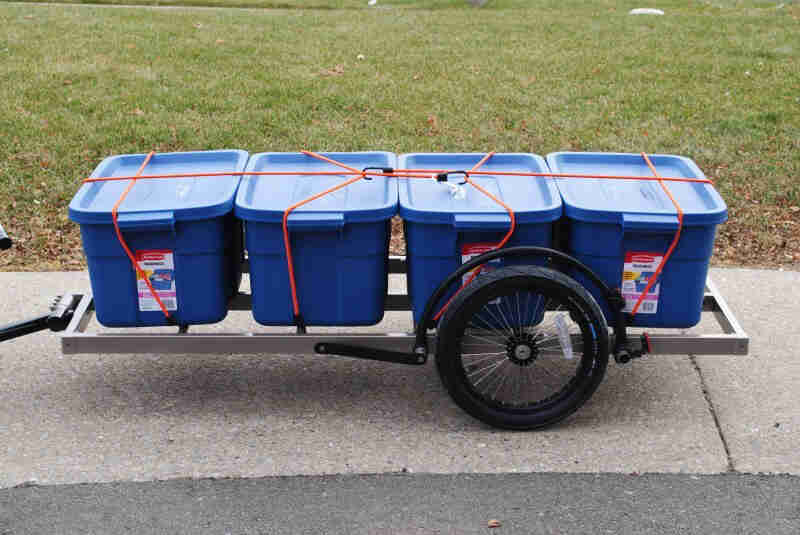 Left side view of a bike trailer, loaded with blue totes, parked on a sidewalk