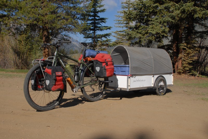 Left side view of a green Surly Ogre bike, with gear and a covered trailer, on a dirt lot with pine trees behind it