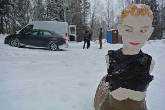 Front view of a Deer Boy statue, in deep snow on a parking lot, with a couple of vehicles and people in the background
