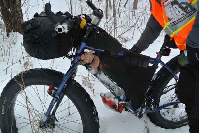 Right side view of a blue Surly fat bike, loaded with gear, standing in snow with a cyclist holding the top tube