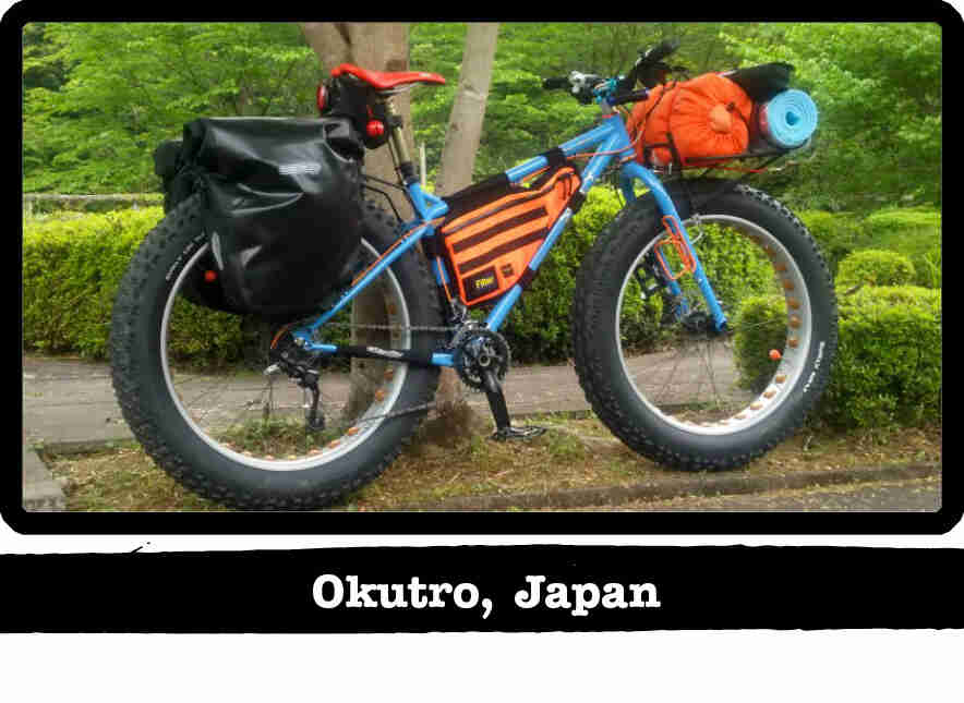 Right side view of a Surly fat bike, blue, loaded with gear, in front of green bushes - Okutro, Japan tag below image