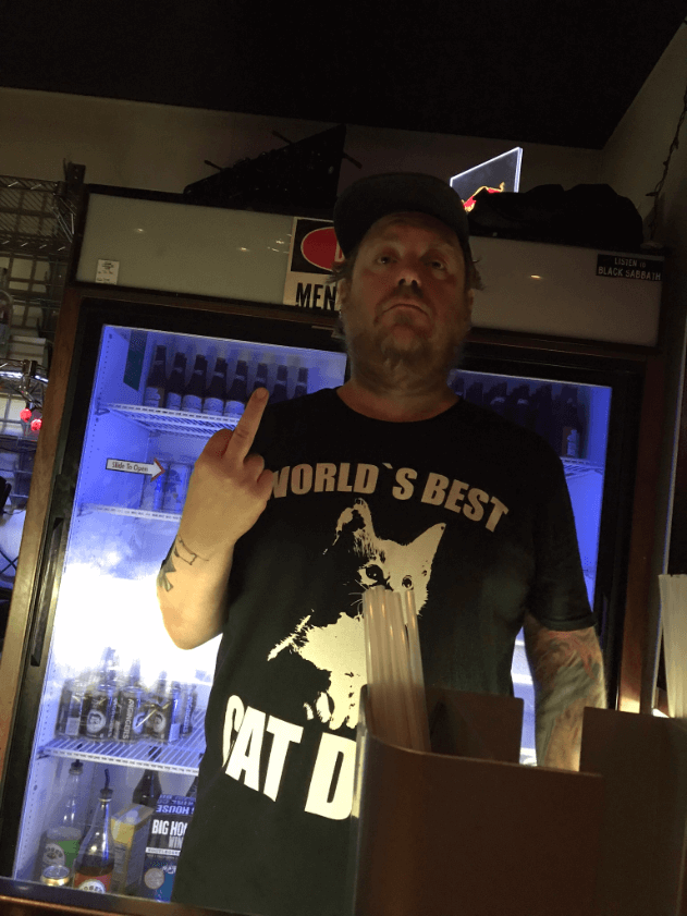 Front view of a person wearing a black t-shirt and showing their middle finger, standing behind a dim lit bar