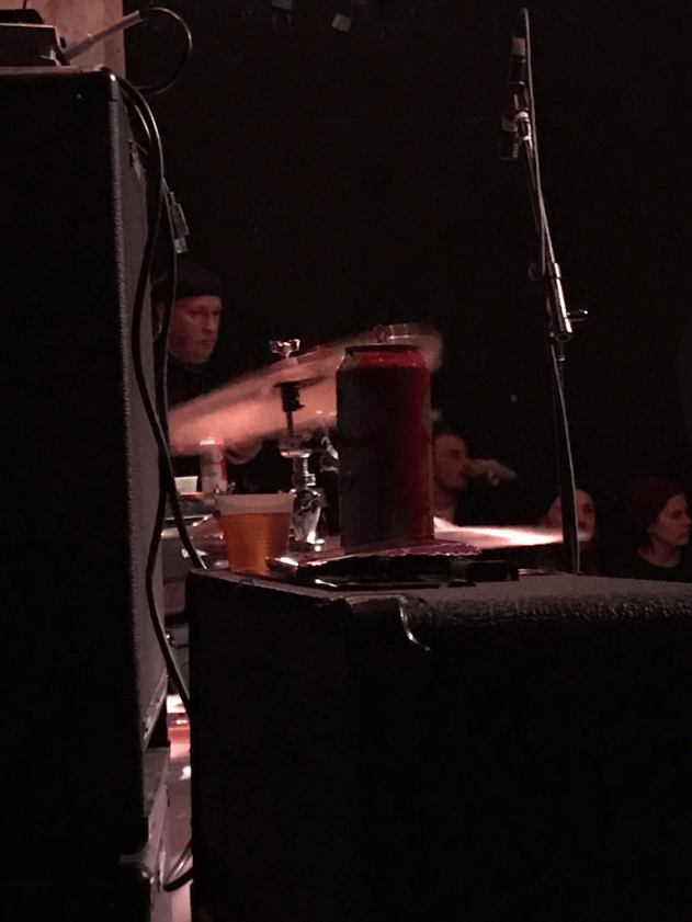 Two speakers with one having a couple of beers on top with a drummer and drum set in the background