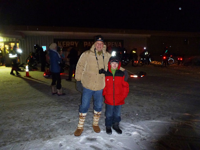 Front view of an adult and a child, standing side by side in front of a building, on a snow covered parking lot at night