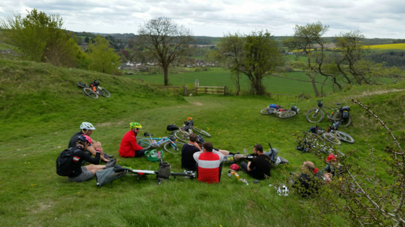 A group of cyclist sitting on a grassy hills with their bikes, with trees and hills in the background