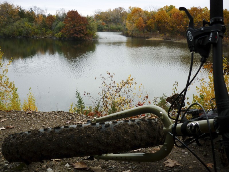 Front end view of a green Surly fat bike, laying on a dirt bank with a pond below, and changing trees in the background