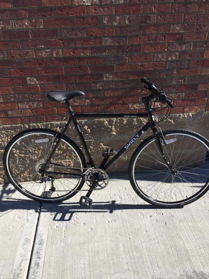 Right side view of a black Surly Cross Check bike, parked on a sidewalk against a red brick wall