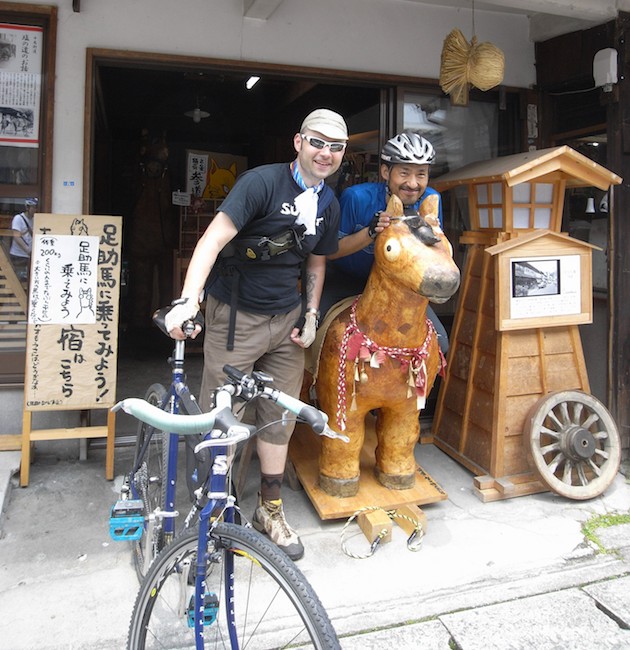 A cyclist standing with a Surly Travelers Check bike, blue and a person standing behind a wooden horse at a store front 