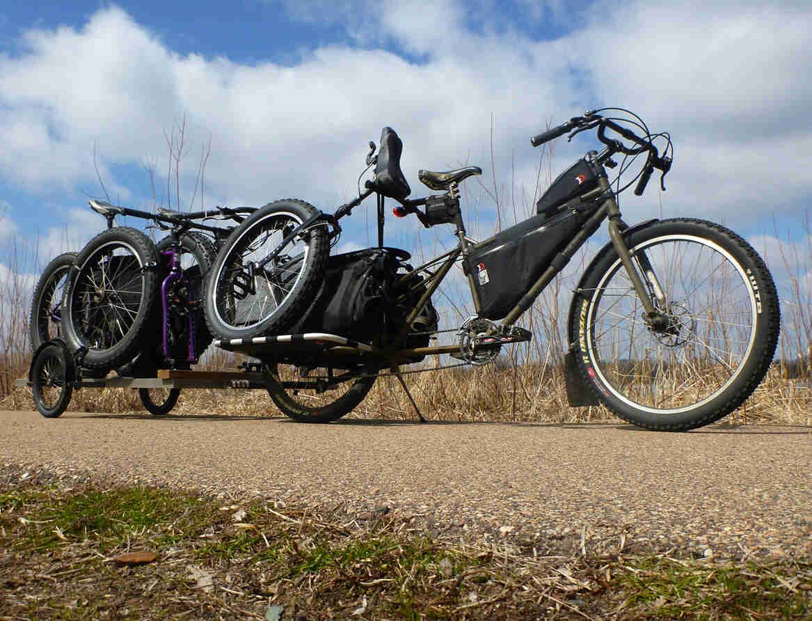 Right side, ground level view of a Surly Big Dummy bike, with a unicycle on back, and loaded trailer hitched behind