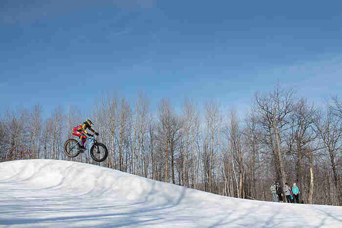 Right side view of a cyclist going airborne on a fat bike at a ski hill, with trees and blue sky in the background