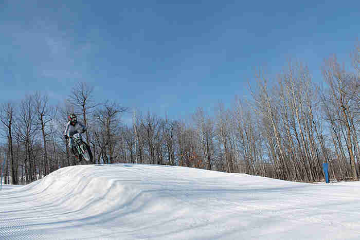 Front view of a cyclist on a fat bike, going airborne off of a berm at a ski hill, with trees in the background