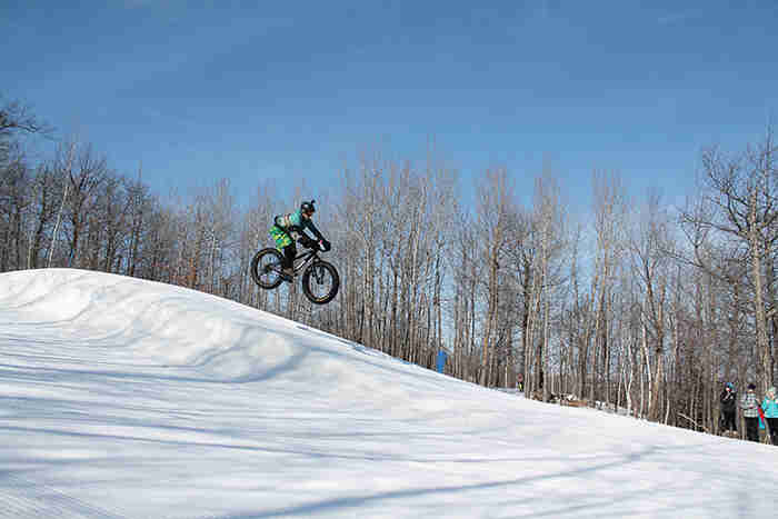 Right side profile view of a cyclist, going airborne on a fat bike at a ski hill, with trees in the background