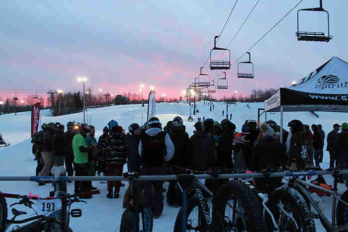Rear view of spectators looking up a ski hill, with ski lift cars above, at dusk