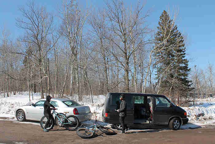 Two cyclists standing on a road next to their bikes beside a van and car, with snowy ground and trees in the background