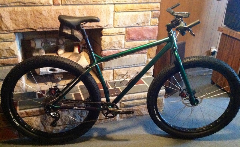 Right side view of a green Surly Krampus bike, leaning on the base of an interior, stone fireplace