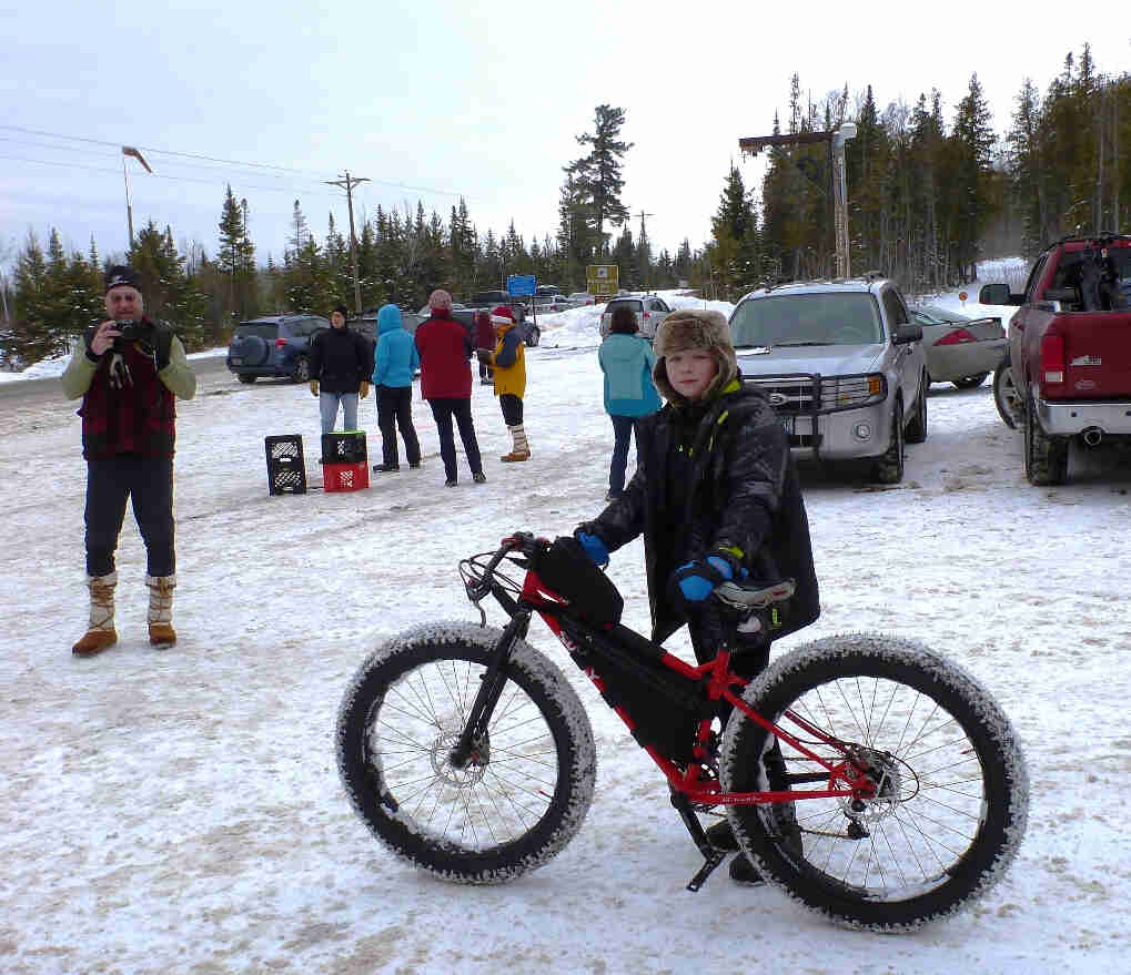 Left side view of a red Surly fat bike, with a kid standing on the right side, on a snow covered parking lot