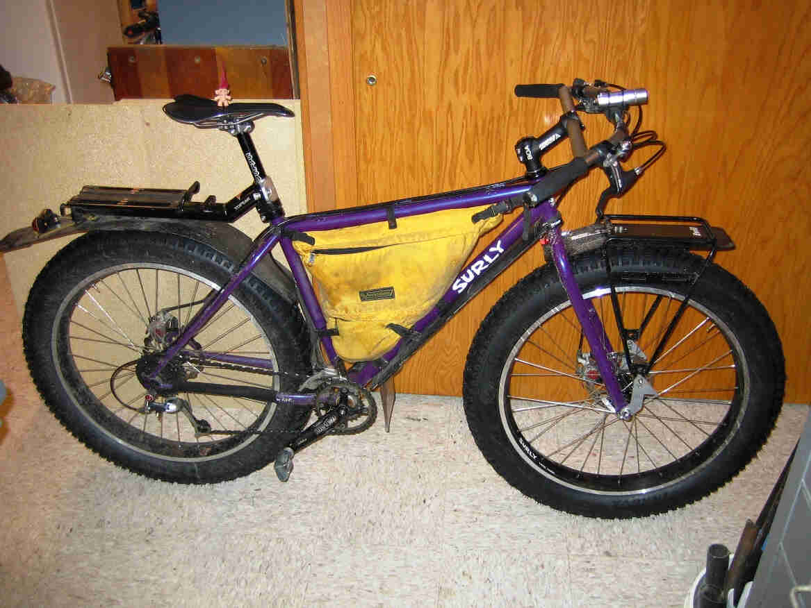 Right side view of a purple Surly Pugsley fat bike with frame pack, leaning against a wood wall in a room