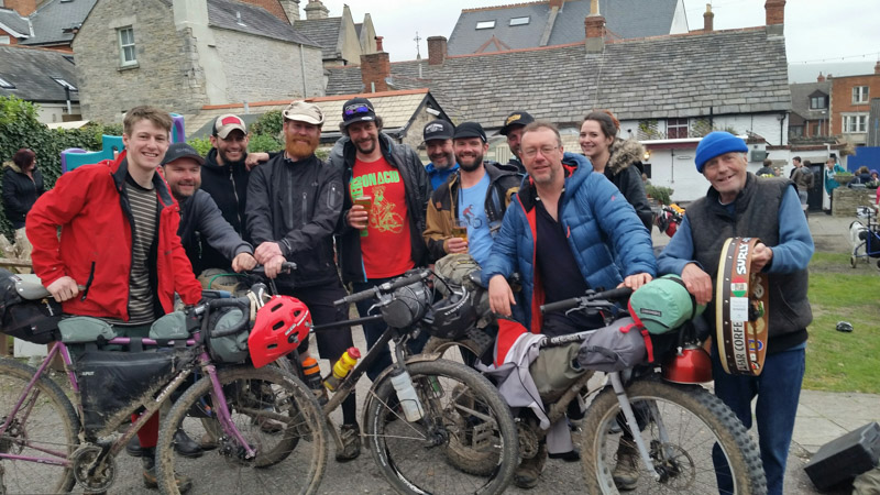 Front view of cyclists posing with their bikes, with village buildings in the background