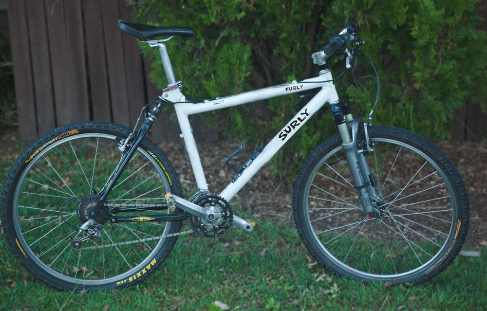 Right side view of a white full suspension bike, parked in grass in front of a wood fence