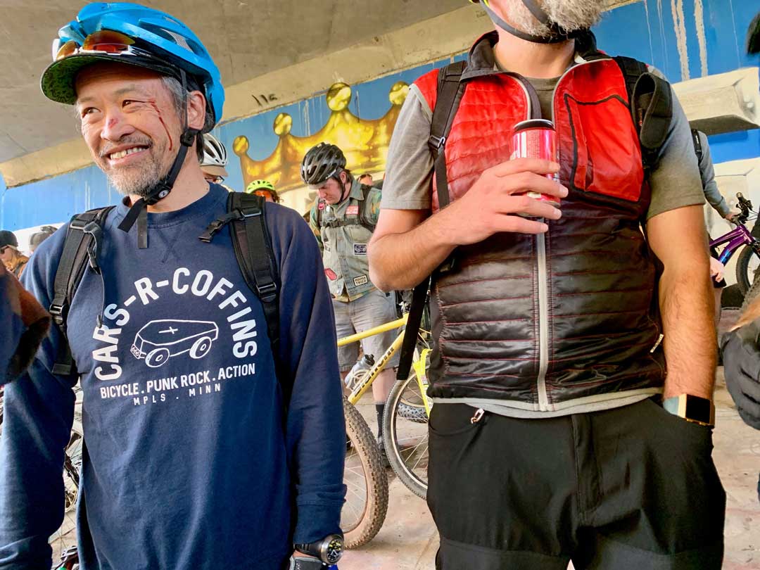 Smiling person with blood near their left eye wearing a blue bike helmet stands next to a person holding a red beer can