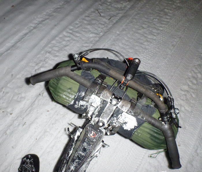 Downward view of a handlebar with a headlight and gear pack, on a bike that's standing on snow at night