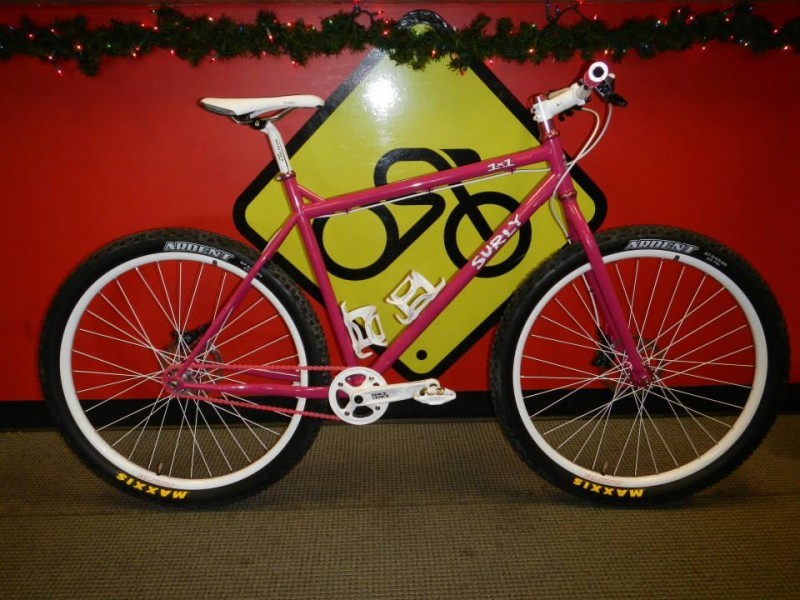 Right side view of a pink Surly 1x1 bike, parked in front of a red wall, with a yellow biking sign painted on it