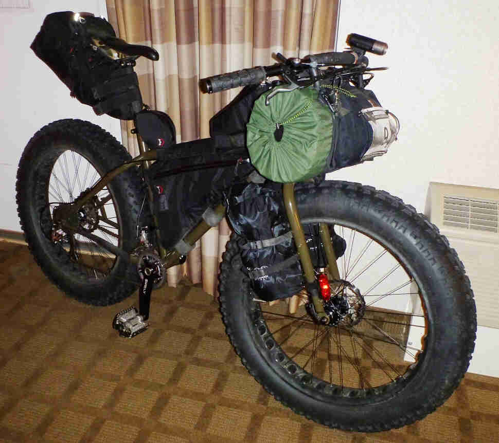 Right side view of an olive drab Surly Moonlander fat bike, loaded with gear, leaning against a wall in a hotel room