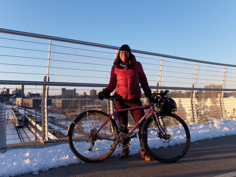 Right side view of a purple Surly Straggler bike, with cyclist standing behind, on a bike bridge with snow on the sides