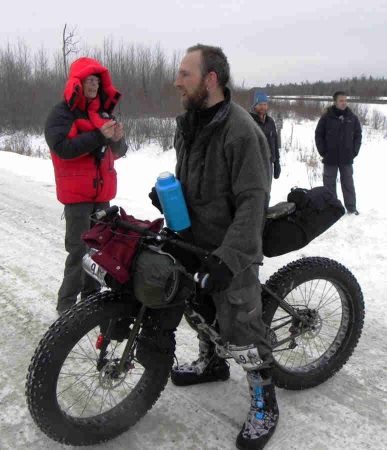 Left side view of a Surly Moonlander fat bike, loaded with gear and a cyclist standing over, on an icy gravel road