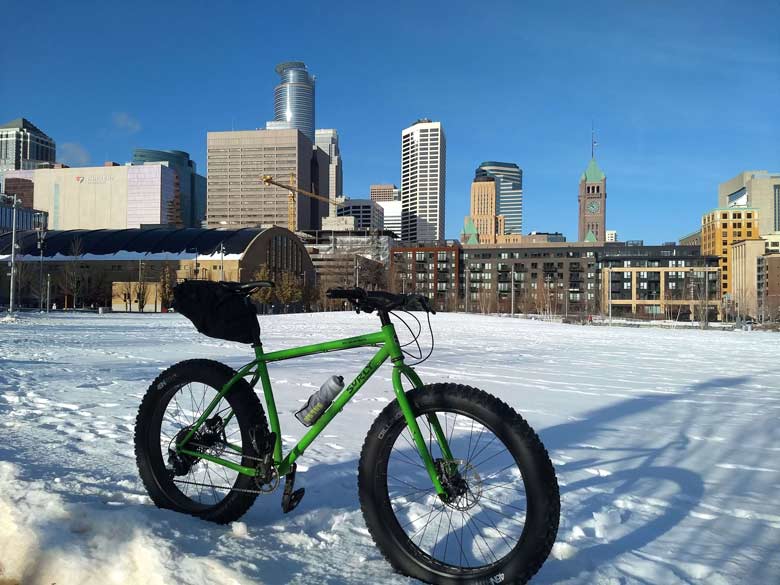 Surly Ice Cream Truck fat bike,green, with seat pack and water bottle in snowy field and buildings of Minneapolis behind